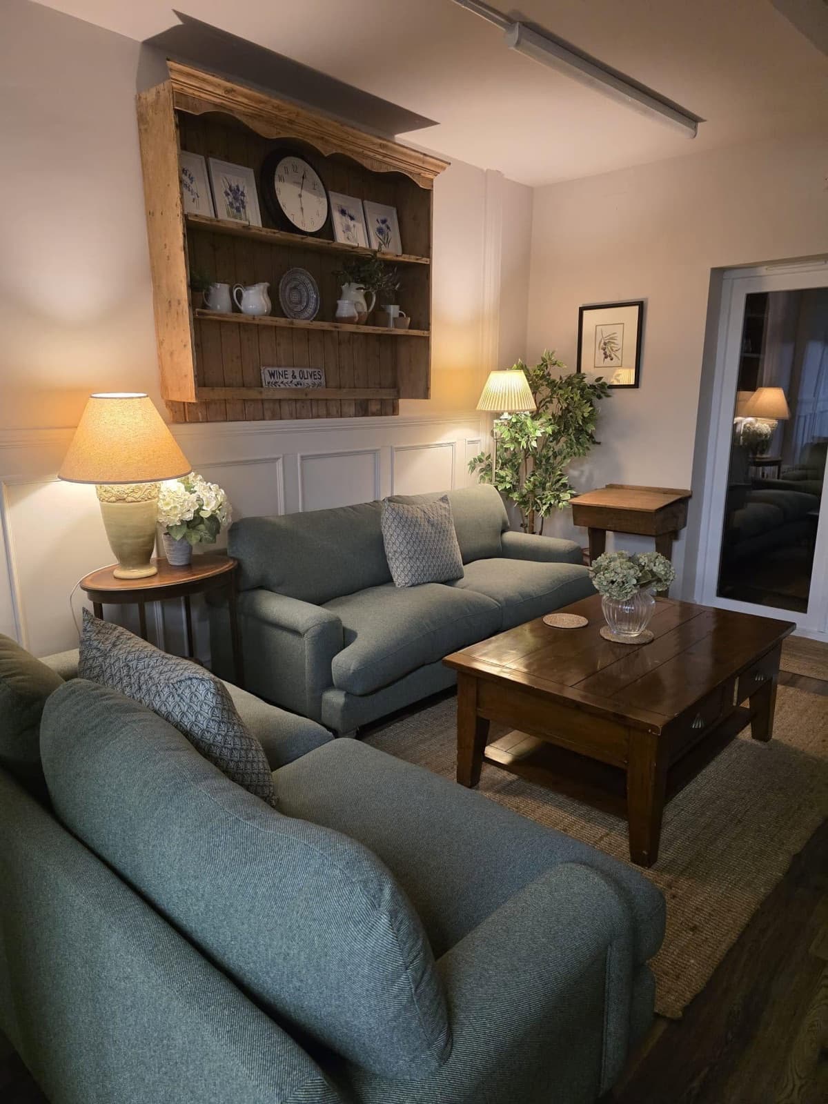 The snug at The Olive Tree — sage green sofas, warm lamplight and a dresser of crockery